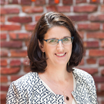 Close-up of Lynette in a black and white patterned jacket in front of a red brick wall.