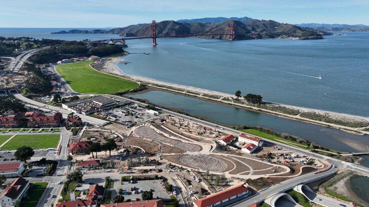 Photograph of present-day Presidio Parkway. Includes overhead view of buildings, streets, greenery, and construction around Battery Bluff park. Golden gate bridge, clear blue skies, the Bay, and mountains in the background can also be seen.
