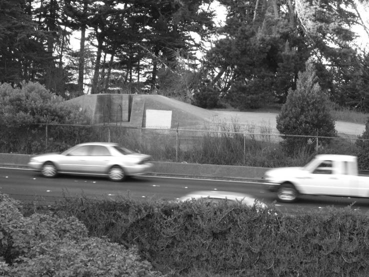 Greyscale photograph. Includes two sedans and a truck in motion on a street, and various trees in the background and foreground. 