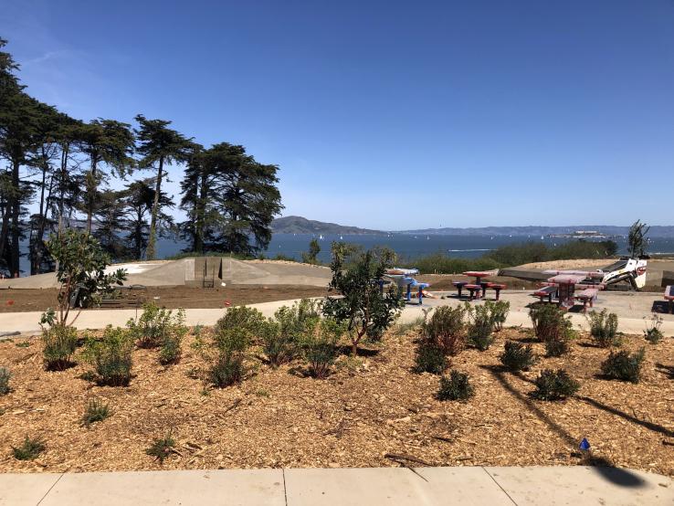 Photograph of present-day Battery Bluff. Includes dirt terrain, picnic tables, a sidewalk, sparse greenery, and mountains in the background. Skies are blue and clear.