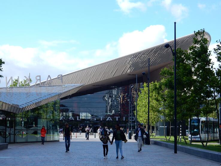 People walking into Rotterdam Central Station on a semi-sunny day. Trees line the concrete path toward the station.
