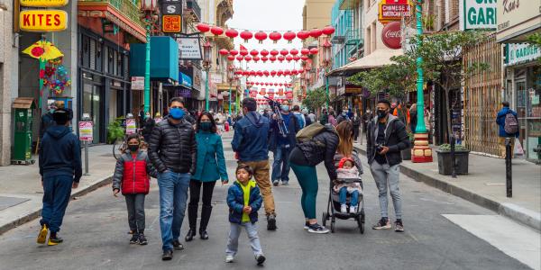 Chinatown, San Francisco street during a cloudy day. Masked children and adult pedestrians walk on the street. Sidewalks are surrounded by small storefronts and signs. Red lanterns hang from the buildings above the street.