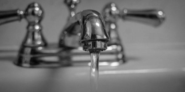 Black and white photo of a running water faucet. Faucet head is in focus, handles and sink are blurry.