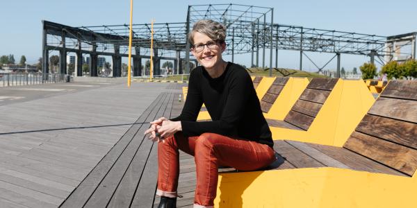 Karen Steen wearing long red pants, a black long-sleeved shirt, and black boots. Karen is sitting on yellow wooden benches outside at Brooklyn Basin on a sunny day.