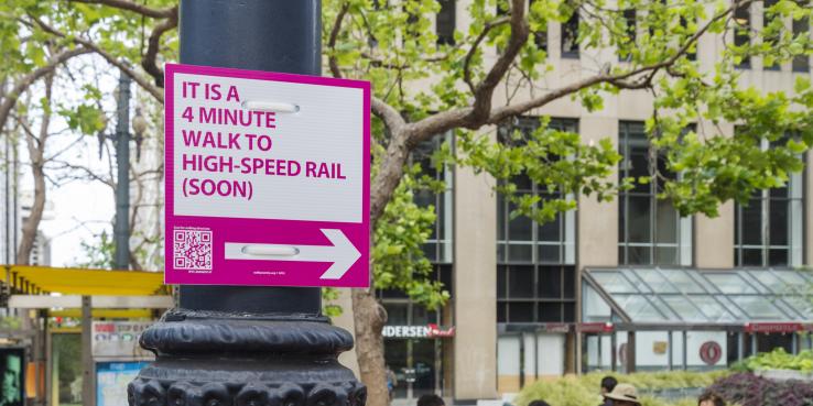 Pink and white sign strapped to a lightpost on a city street. Sign states "It is a 4 minute walk to high-speed rail (soon)" with a QR code and an arrow pointing right. In the background, there are trees, a yellow bus stop, and a tall, tan building with several tall windows.