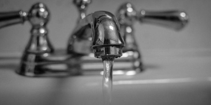 Black and white photo of a running water faucet. Faucet head is in focus, handles and sink are blurry.