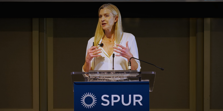 SPUR President and CEO Alicia John-Baptiste speaking at a podium with the SPUR logo in white on a blue background