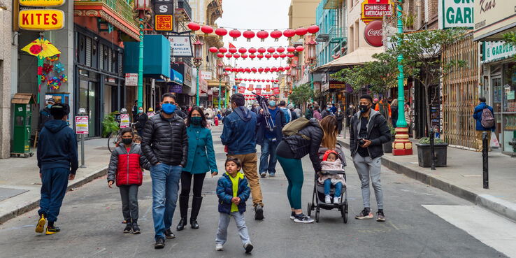 Chinatown, San Francisco street during a cloudy day. Masked children and adult pedestrians walk on the street. Sidewalks are surrounded by small storefronts and signs. Red lanterns hang from the buildings above the street.
