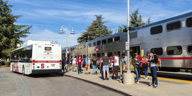 People waiting outside the Palo Alto Caltrain station. Caltrain and Bus are present. Lightpost with two lamps next to the people.
