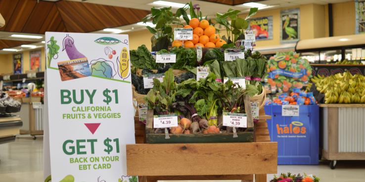 Grocery store stand with fruits and vegetables. Sign on the left side describing the benefits of the EBT pilot program