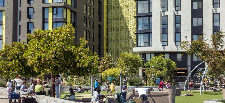 Photograph of San Francisco apartment complex colored in light green, grey, and white. In front of the building, pedestrians walk by and children play on a playground with lots of surrounding greenery.