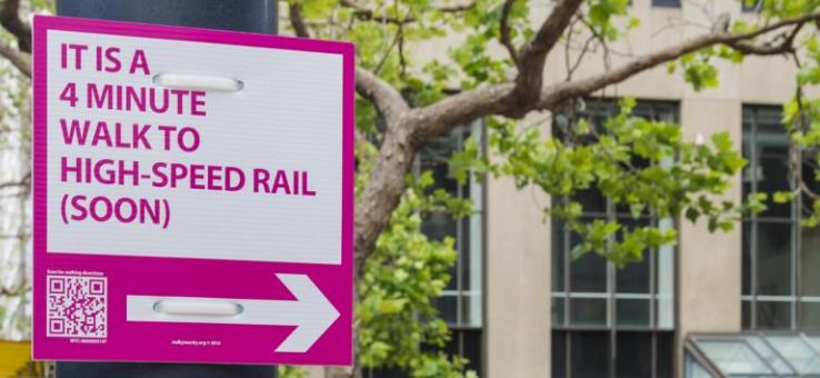 Pink sign that states "it is a 4 minute walk to high-speed rail (soon)" on a city street