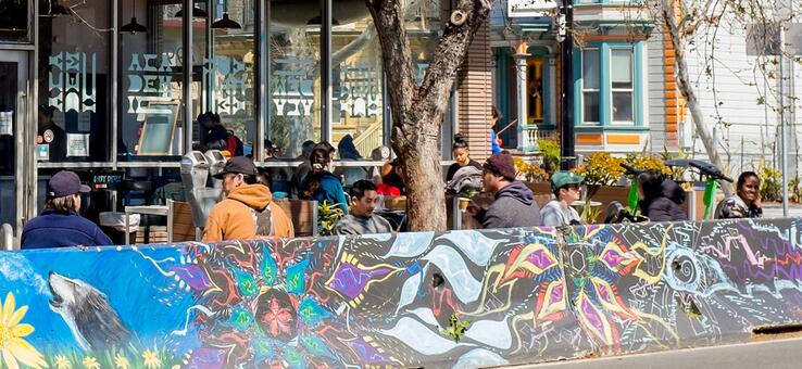 people sitting in a parklet with a colorful mural outside a cafe