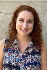 Outdoor grey brick background. Subject is a woman with medium-length curly, red hair wearing a sleeveless, collared patterned shirt.