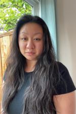 Indoor backdrop next to windows with greenery outside. Subject is a woman with long, black hair and a black t-shirt.