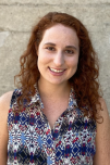 Outdoor grey brick background. Subject is a woman with medium-length curly, red hair wearing a sleeveless, collared patterned shirt.